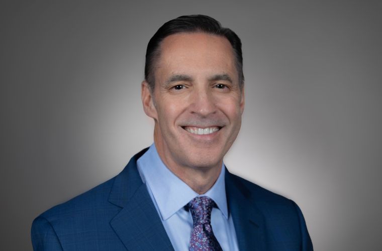 A headshot of Garrick "Gary" Richards smiling. He is wearing a navy suit jacket, with a light blue collared shirt underneath and a floral pattern tie. He has short dark hair.