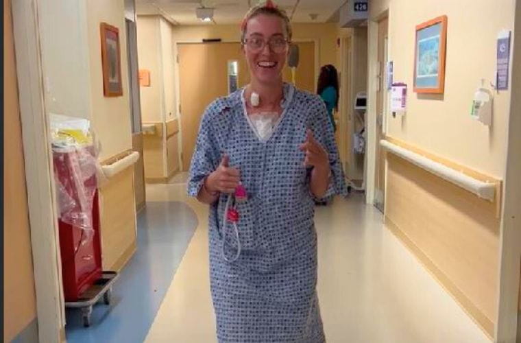 A blonde woman in a blue hospital gown walks unassisted.