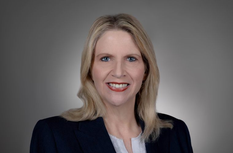 A headshot of Ann Marie Swindler smiling. She has below shoulder length straight blonde hair and is wearing a black jacket with a white shirt underneath. 