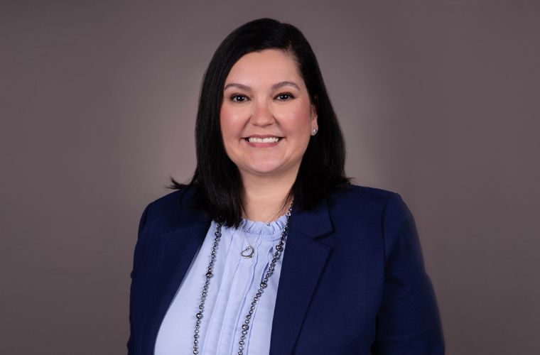 A headshot of a young woman smiling. She has dark shoulder straight hair and is wearing a navy blazer with a light blue shirt underneath, a long, beaded sliver necklace and a shorter heart-shaped silver necklace.