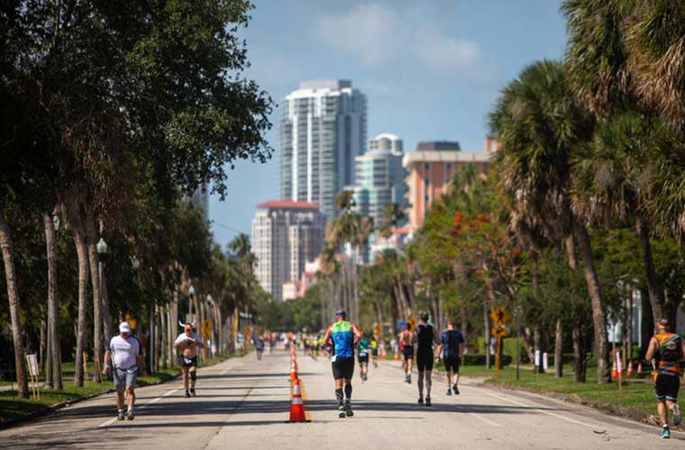A group of people dressed in athletic clothing run on a scenic street lined with trees.