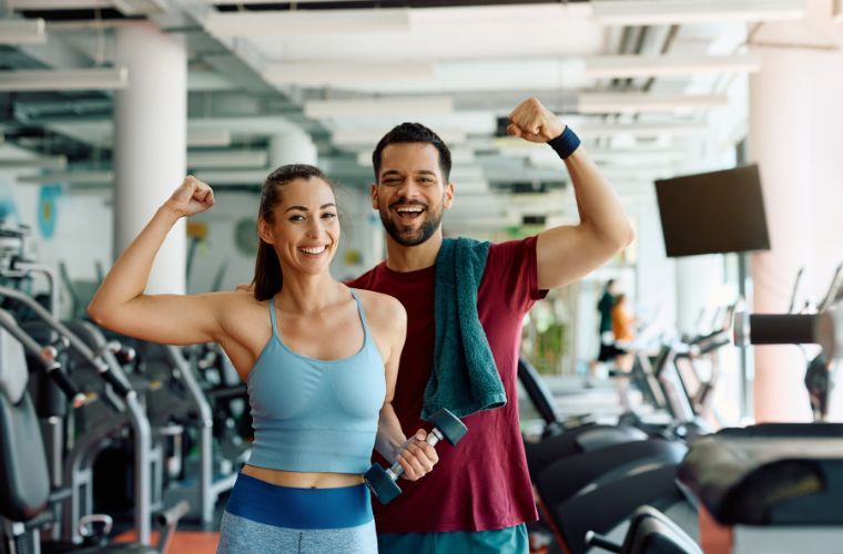 A young woman (left) wearing a blue spandex fitted top and matching pants stands next to a young man (right) with a maroon shirt and blue shorts with a blue towel thrown over his left shoulder. They are both making a flexing gesture with one arm and standing inside a gym in front of equipment. 