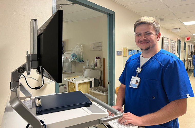 A young man dressed in hospital blue scrubs looks at a computer as he stands in front of a hospital room.