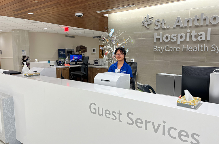 A young woman dressed in blue polo shirt stands behind a desk that says guest services in the lobby of a building. 
