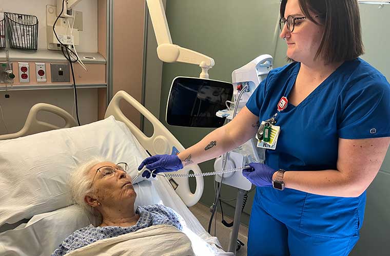 A woman dressed in blue hospital scrubs takes the temperature of a woman with gray hair and wearing glasses lying in a bed. 