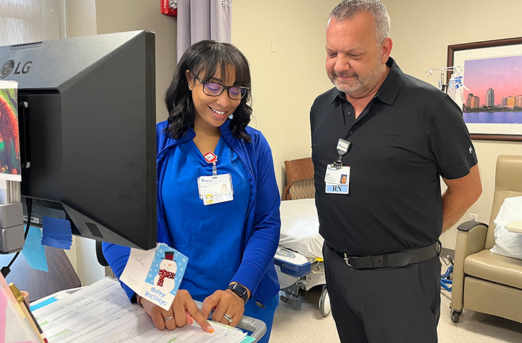 A woman dressed in blue hospital scrubs talks with a man dressed all in black as they stand at a computer and look at a report on the computer cart.