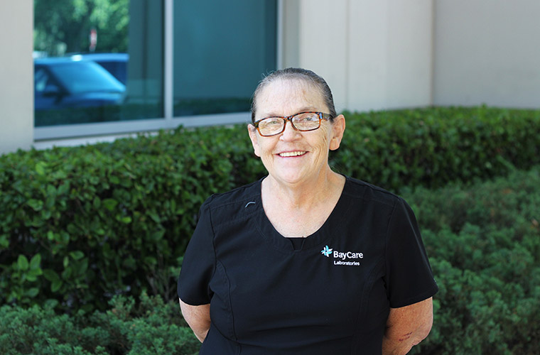 A middle-aged woman smiles for a photo outdoors while wearing black scrubs with the words "BayCare Laboratories" stitched on them. The woman is standing in front of some shrubs, outside of an office building.