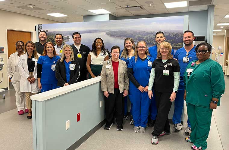 A group of people, some dressed in regular street clothes while others are dressed in hospital scrubs, smile as they stand in a large room.