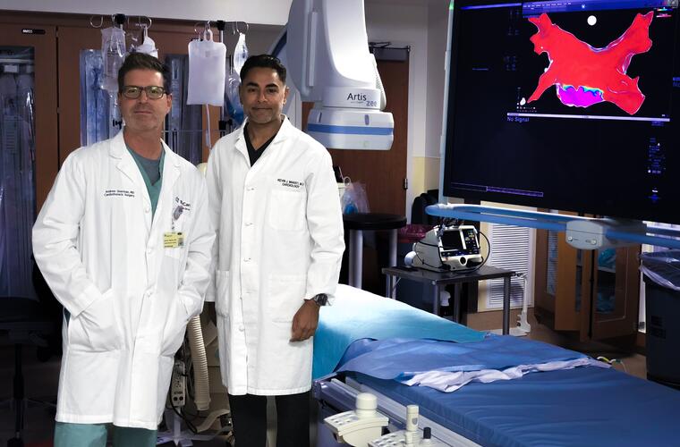 Two men in lab coats stand in front of an operation table with a large TV screen showing a mapping image.
