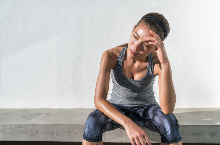 A woman in a grey tank top and cropped workout pants is sitting on a bench with her head in her hands, overwhelmed by the hot temperature outside.