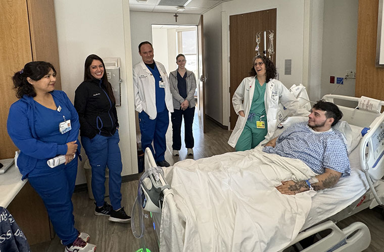 A group of men and women dressed in hospital scrubs and some in doctor’s coats surround a young man lying in a hospital bed.
