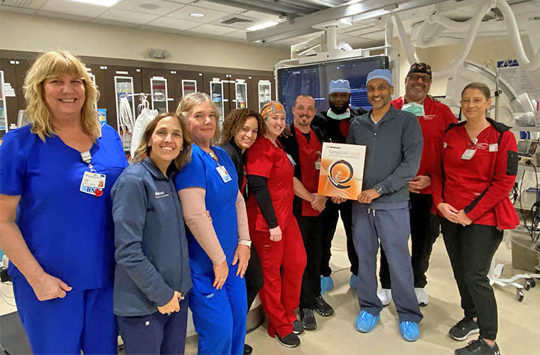 Ten healthcare professionals smile as they stand in a procedure room holding a box that says Symplciity Spyral.