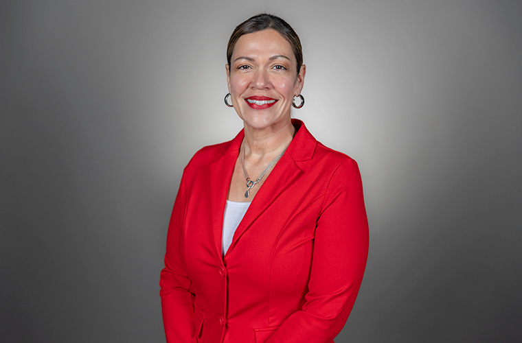 A headshot of Maya Perez.  She has dark hair pulled back, with silver earrings and necklace in a red jacket and she is smiling. 