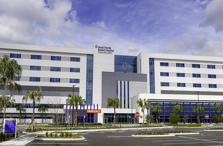 The exterior of South Florida Baptist Hospital. It’s a large hospital building against a blue sky with white clouds. Palm trees line the front of the building.