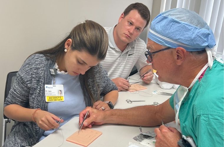 An older male surgeon wearing blue scrubs is across a classroom table from a female and a male resident. They all hold small instruments and sewing supplies are on the table.