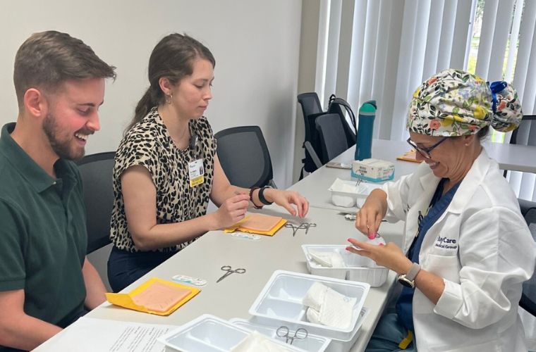 A female surgeon wearing a lab coat smiles while sitting across a classroom table from a male and a female resident.  Surgical sewing supplies are on the table.