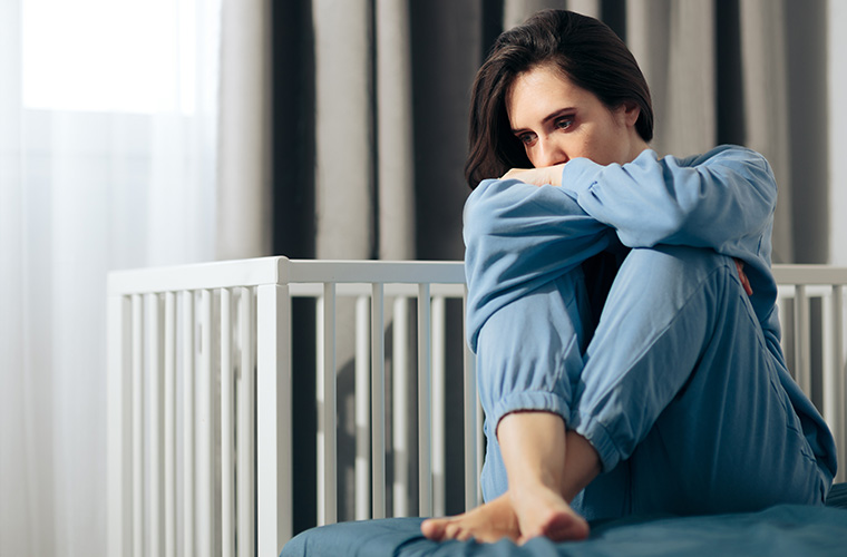 A woman sits on floor next to a baby crib and has her head propped up on her crossed arms.
