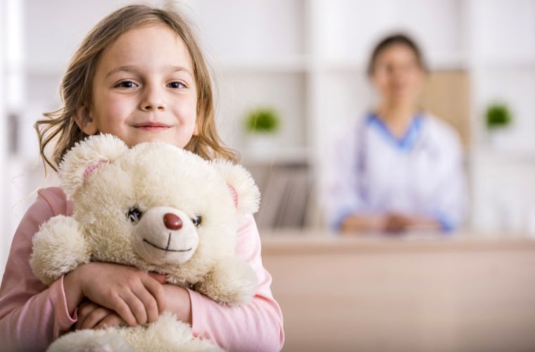 A school-aged young blonde girl hugs a cream teddy bear while a dark-haired female clinician smiles at her from behind a workstation.
