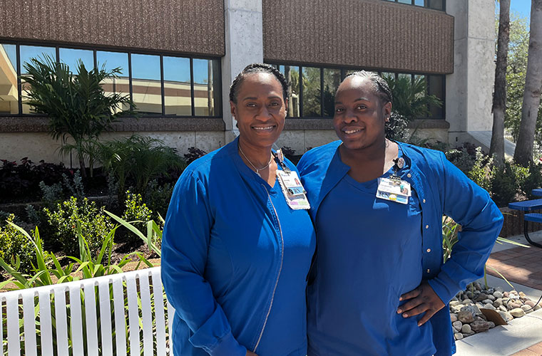 Two Black women dressed in blue scrubs stand arm in arm outside a building.