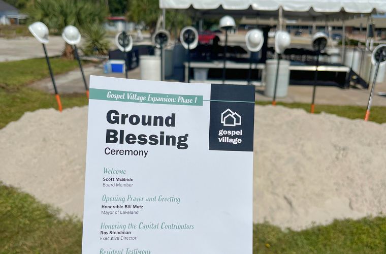 A white flyer for a groundbreaking ceremony is held up in front of the site where the ceremony took place, marked by multiple shovels in the ground.