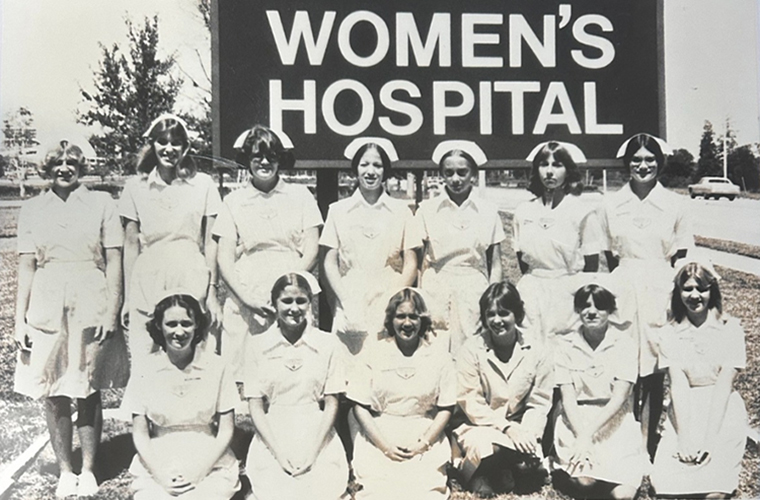 A black and white image of two rows of nurses wearing white caps and white dresses. They are in front of a sign that says Women's Hospital.