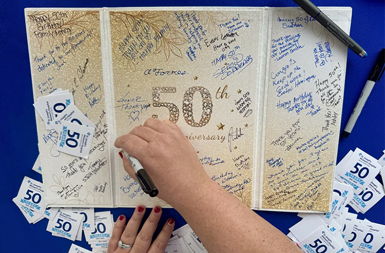 A hand writing with a black marker is adding a signature on a large card that says 50th Anniversary.