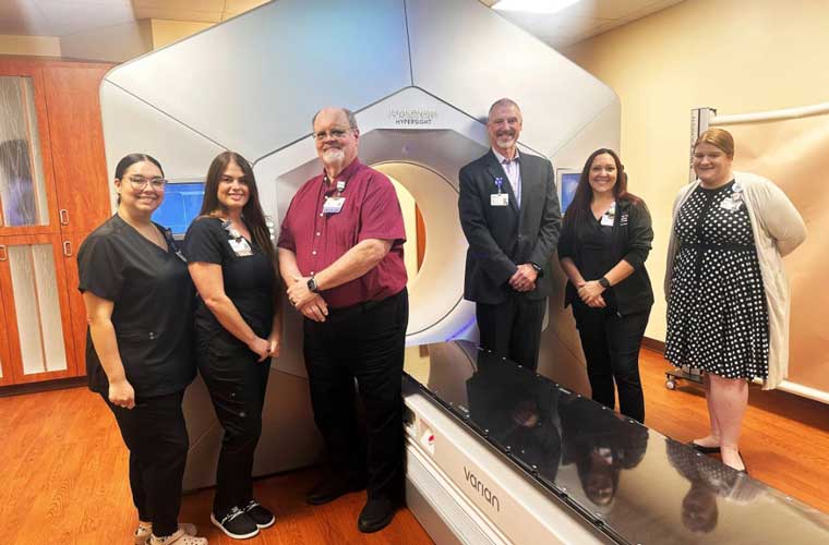 A group of people – some dressed in hospital scrubs – stand around a new piece of equipment in a room. 