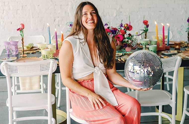 A woman sits in front of a table set with dinnerware and candles as she holds a mirrored ball.