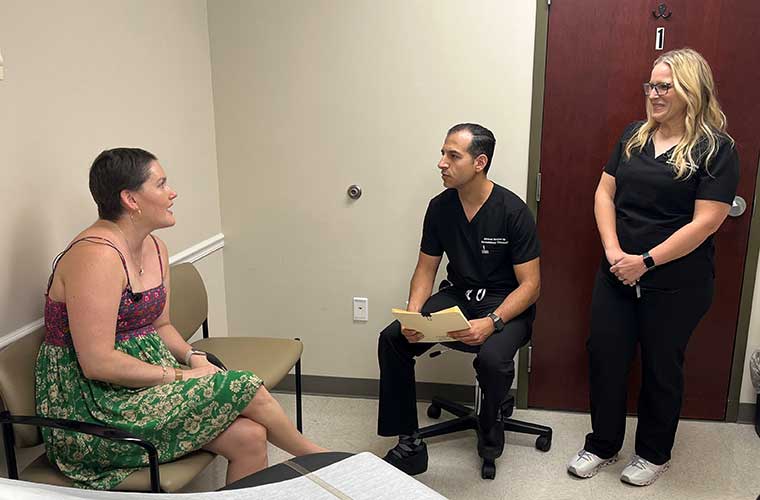 A woman with a short haircut dressed in mini dress talks with a man who is sitting and a woman who is standing and they are both dressed in black hospital scrubs.
