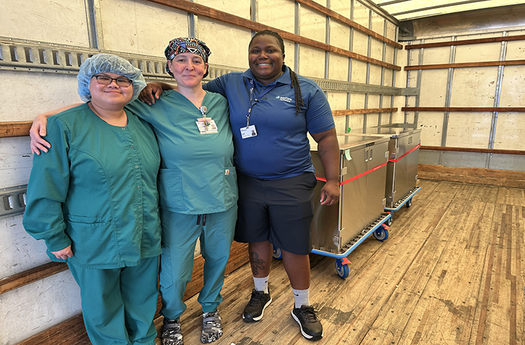 Three hospital staff members, two in medical scrubs and one in a service uniform, smiling in the back of delivery truck loaded with two, large metal carts.