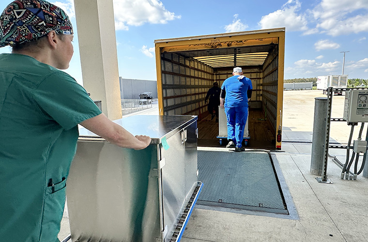 Two medical professionals push large metal carts into the back of an open delivery truck at a hospital loading dock.