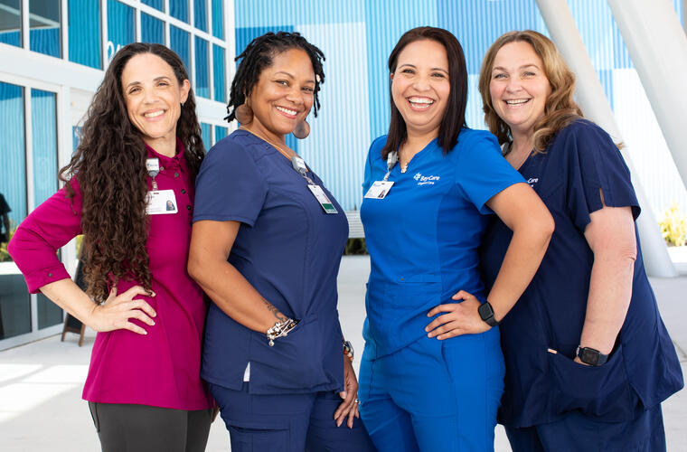 Four female team members in different color scrubs pose smiling in front of a BayCare Health System building.