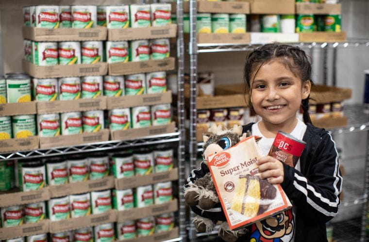 A young girl stands in front of shelves, holding a variety boxed and canned food with a cheerful expression on her face.