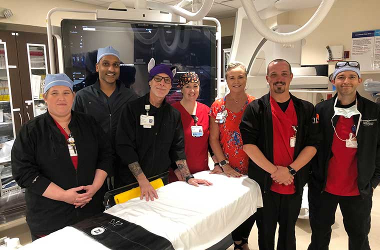 Seven clinicians stand between the big monitor and surgical table in the Cath Lab after a procedure.