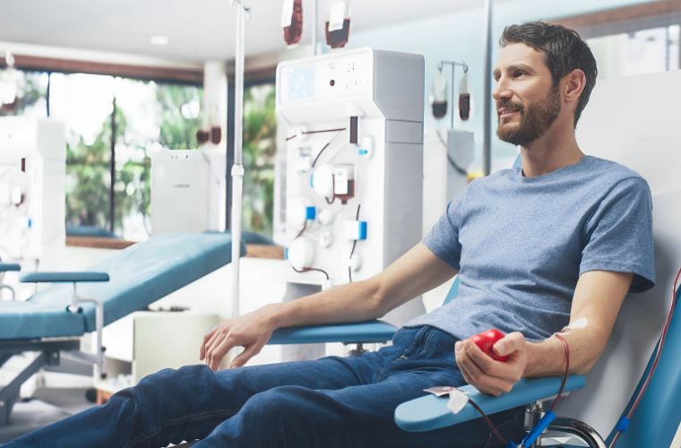 A man with short hair and a beard wearing jeans and a light blue T-shirt is lying back in a chair and donating blood.