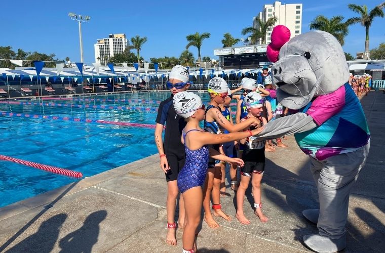 A person dressed in a manatee costume is surrounded by children wearing swimsuits at a pool.