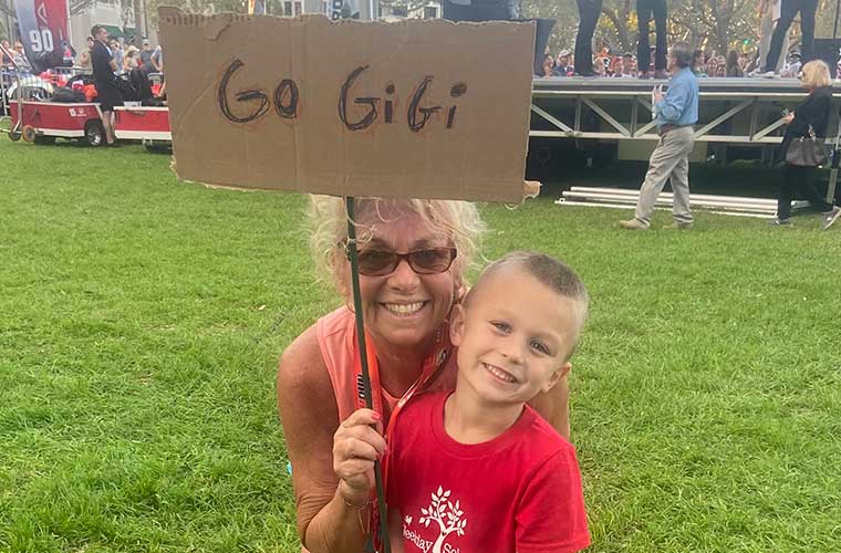 A woman wearing sunglasses and dressed in a tank top hugs a child who is dressed in a red shirt and holding a sign to support someone. 