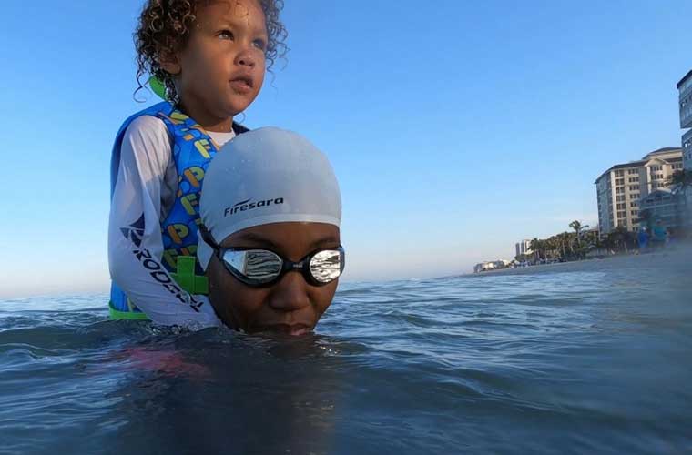 A child dressed in a long-sleeved swim shirt sits on the shoulders of a woman wearing a swim cap and goggles as they both sit in the water.