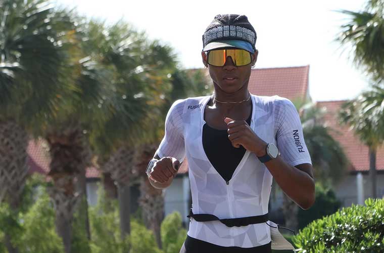 A woman wearing a visor and sunglasses and dressed in athletic clothing runs past homes and palm trees.