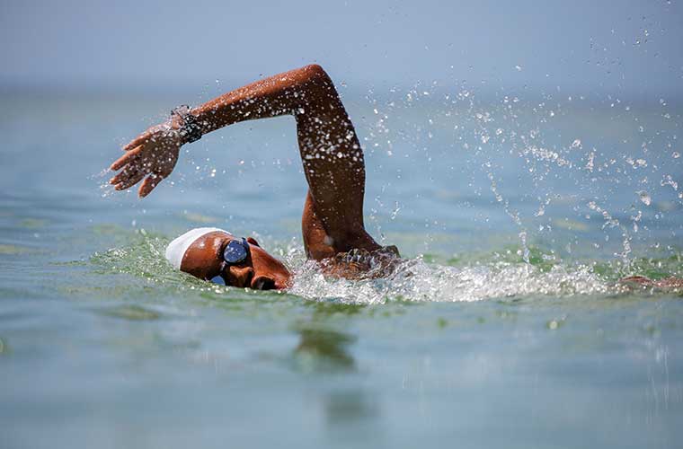 A woman wearing swim goggles and a swim cap raises her left arm as she prepares to take a stroke in the water.