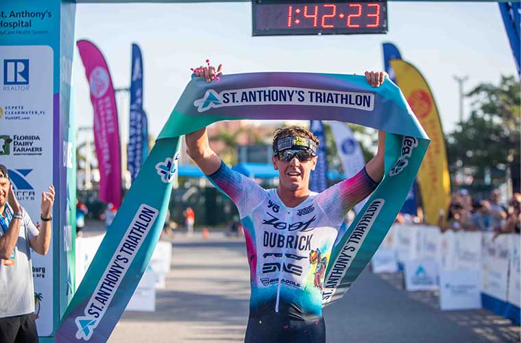A man dressed in athletic clothing holds up the tape at a finish line at an athletic competition.