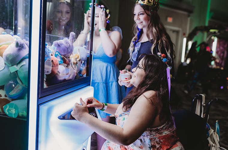 Three teenage girls wearing colorful dresses laughing in front of an arcade machine.