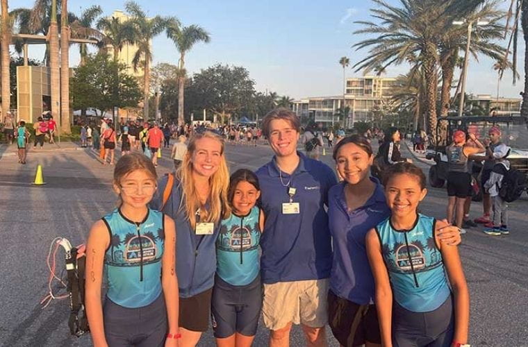 Three children wearing athletic clothing smile for the camera with three adults in a parking lot as they prepare for an athletic event.