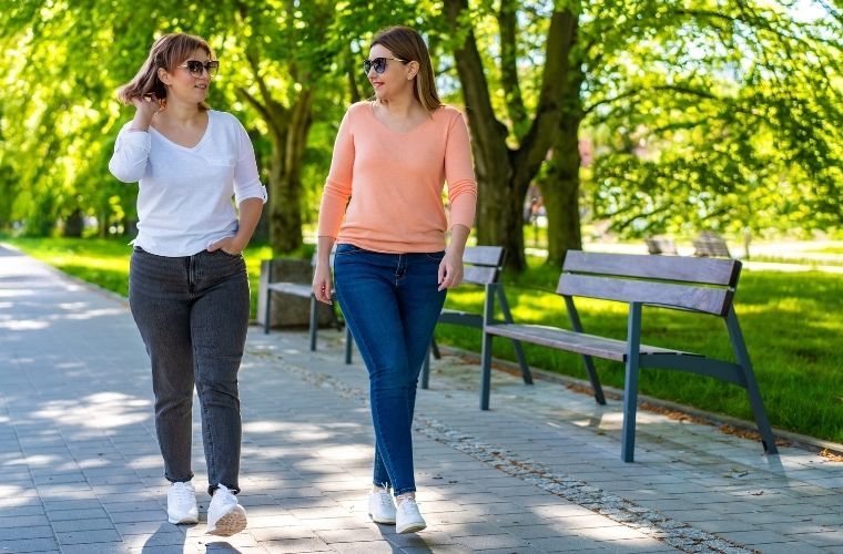 Two women in casual clothes and sunglasses walk together on a sunny, tree-lined park path, talking and enjoying the outdoors.