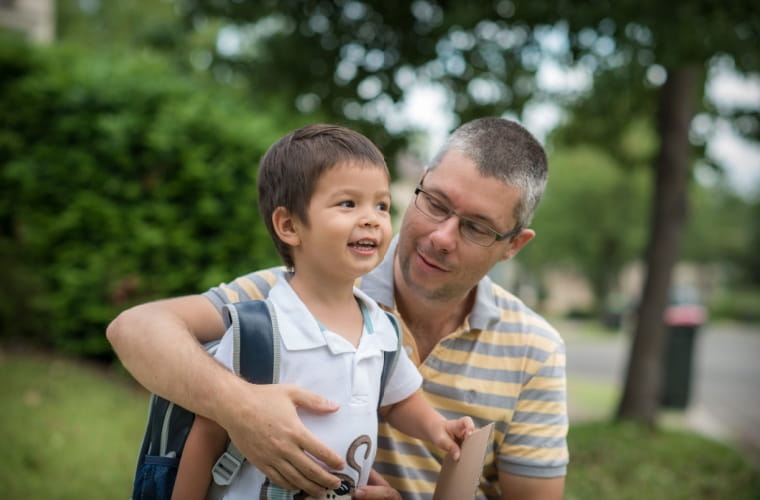 Father helping young son with a backpack get ready for school, both smiling outdoors on a tree-lined sidewalk.