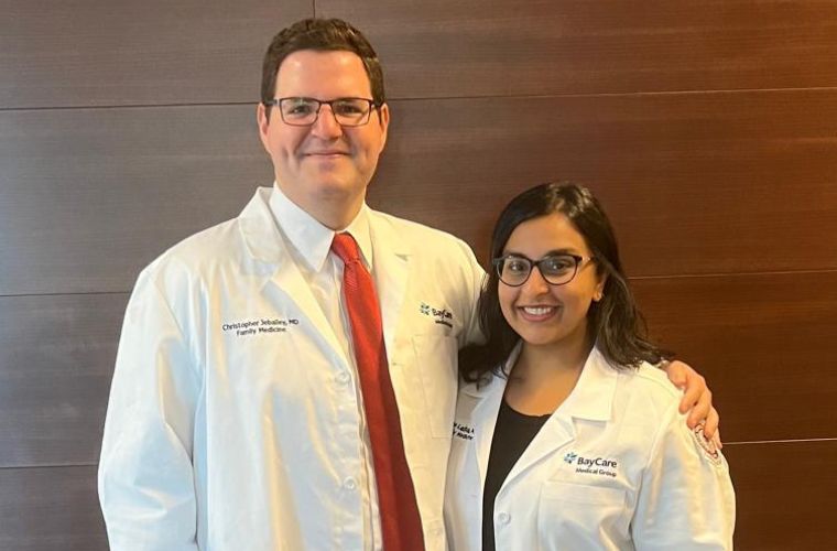 Dr. Christopher Jebailey and Dr. Alina Ladha, both wearing white BayCare medical coats, smile while standing side by side in front of a dark wood wall.