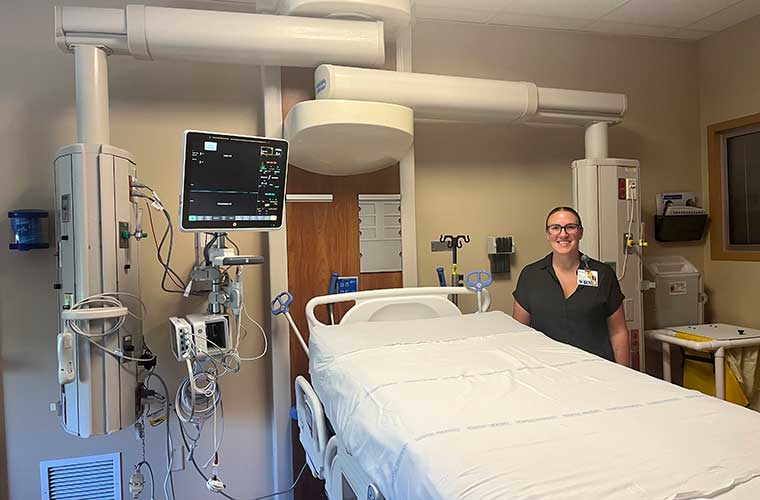 A woman dressed in hospital scrubs stands in a large hospital room surrounded by equipment.