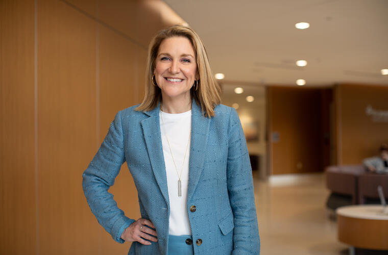 A smiling headshot of Kate Sawa standing indoors in a modern office setting, wearing a textured light blue suit with a white top underneath. She has shoulder-length straight hair and is accessorized with a long pendant necklace. One hand rests on the hip, and the background shows wood-paneled walls, soft seating and warm ambient lighting.