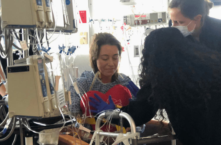 A female patient sits on a chair surrounded by medical equipment as rehab personal help her recover. The patient is holding a stuffed red heart on her chest with a slight grin. 