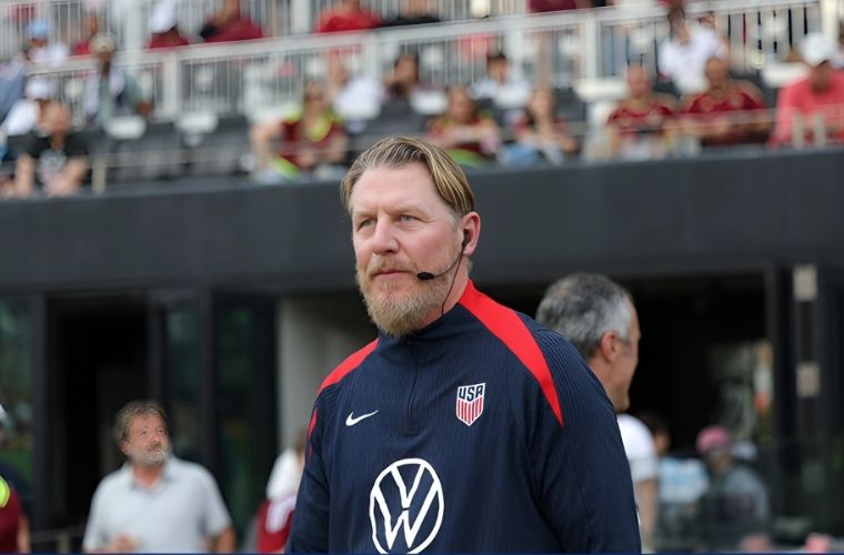 Image of Dr. Kevin Elder; older white male with grey beard, blue eyes, short graying blond hair, wearing a black headset, with an official navy blue American men’s soccer team jacket that features a white Volkswagon logo, white Nike Swoosh logo, red, white and blue USA men’s team official logo, and red stripes from collar to shoulder on each side of the jacket.